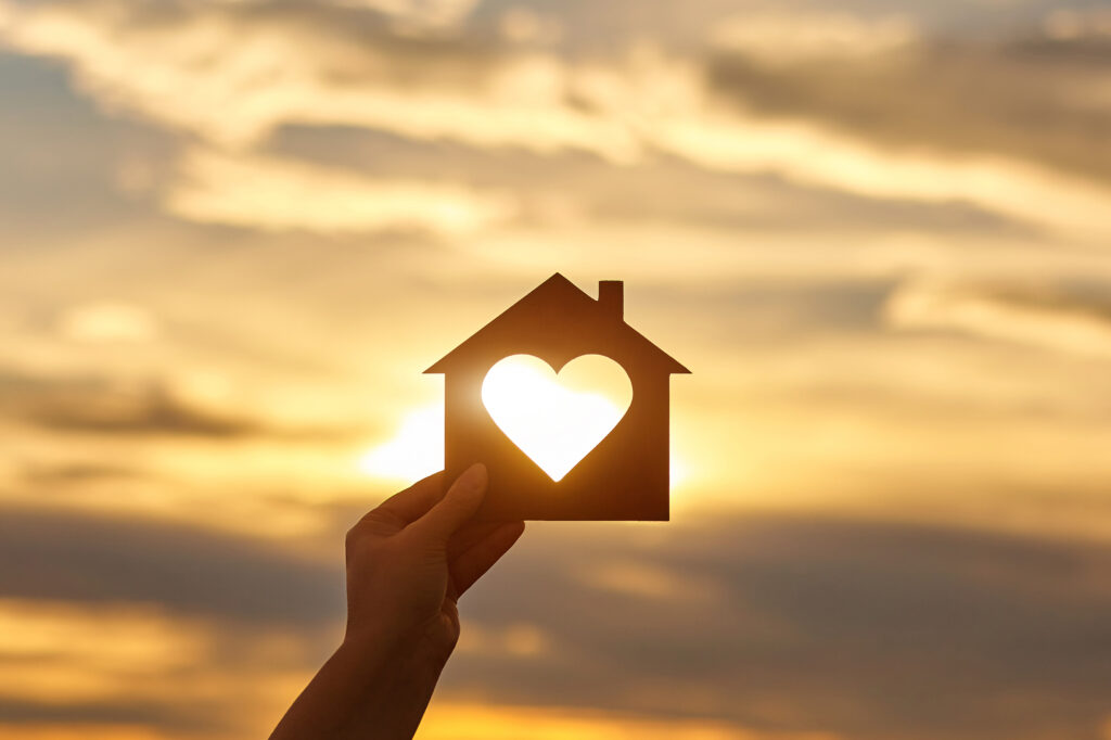 Woman hand holds wooden house in the form of heart against the sun. 