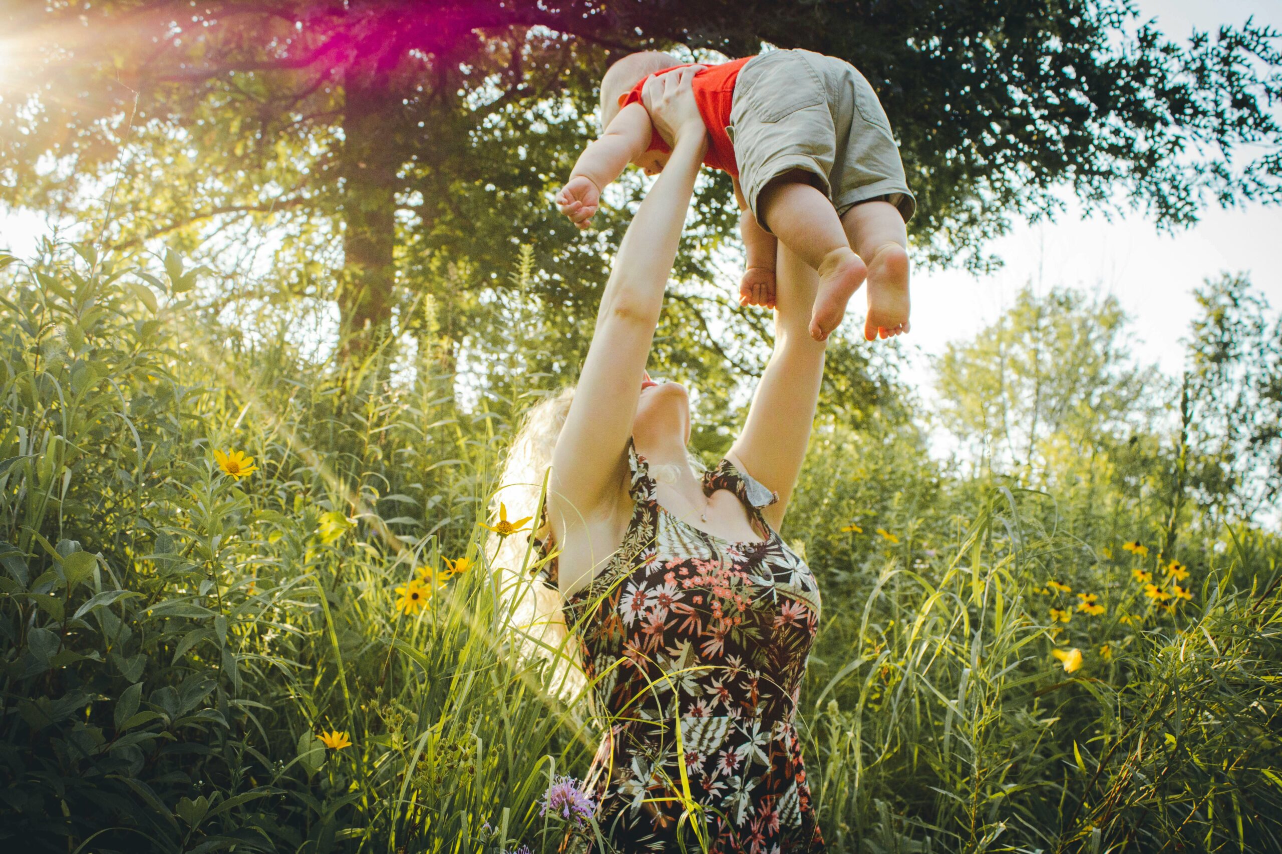 Mother lifting child into the air, surrounded by grass and flowers.